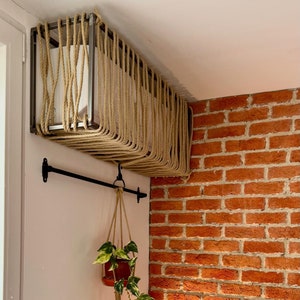 Pode incluir: Um vaso branco com um design de corda está suspenso do teto. O vaso está pendurado em um canto de uma sala com uma parede de tijolo. Uma planta suspensa com folhas verdes está pendurada em uma barra de metal preta.
