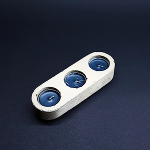 Puede incluir: Un portavelas rectangular de hormigón blanquecino con tres hendiduras circulares, cada una con una vela de té azul oscuro. El soporte está sobre un fondo oscuro, resaltando el diseño minimalista.