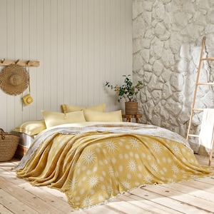 May include: A bed with a yellow and white patterned blanket and pillows. The blanket has a starburst design. The room has a wooden floor and a stone wall. A straw hat and a camera hang on the wall.