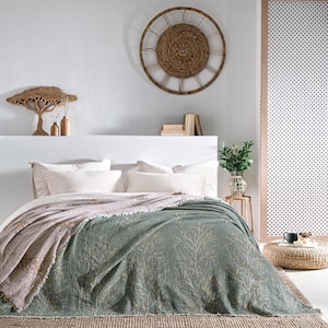 May include: A green and beige patterned throw blanket with a fringed edge, folded on a bed with white pillows. The blanket is on a bed with a white headboard and a woven rug.