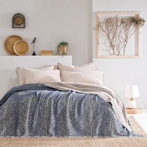 May include: A blue and white patterned throw blanket with a fringed edge, folded on a bed with two beige pillows. The blanket features a design of stylized trees.