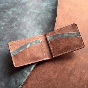 May include: Open brown leather wallet with card slots. The wallet is made of a rich, textured leather and features a contrasting gray interior. The stitching is visible around the edges. The wallet is displayed open, showcasing its interior design.