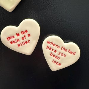 May include: Two white heart-shaped magnets with red text. One magnet reads "this is the skin of a killer" and the other reads "where the hell have you been loca". The magnets are on a black surface.