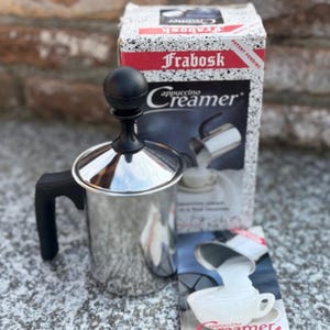Könnte beinhalten: Ein Frabosk Cappuccino-Milchaufschäumer aus Edelstahl mit schwarzem Griff und einem schwarzen, kugelförmigen Knauf auf dem Deckel. Das Produkt wird mit der originalen Kartonverpackung mit Markennamen und einer Bedienungsanleitung geliefert.