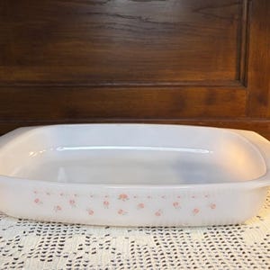 May include: A white, rectangular baking dish with rounded edges and handles. The dish is decorated with small pink floral patterns along the bottom edge. The dish is made of a durable material, perfect for baking.