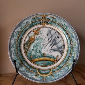 Può includere: Piatto decorativo a tema nautico. Il piatto presenta un'illustrazione dettagliata di una nave in mare, circondata da onde e dettagli di corda nautica. La tavolozza dei colori comprende sfumature di blu, verde e oro.