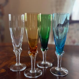Può includere: Un set di quattro flûte da champagne alte con coppe trasparenti, ambra, verdi e blu. Ogni bicchiere presenta uno stelo sottile e testurizzato con un design in vetro intrecciato alla base della coppa e una base circolare in vetro trasparente.