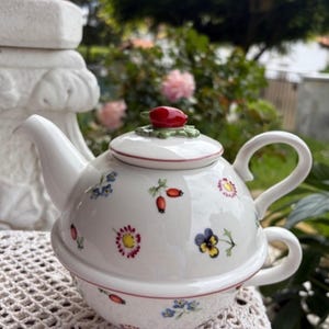 May include: White ceramic teapot with a matching teacup, adorned with colorful floral and berry designs. The lid features a red berry-shaped knob. The teapot is set on a white lace surface, with a blurred green background.