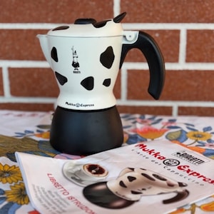 Pode incluir: Uma cafeteira Bialetti Mukka Express com corpo superior branco com manchas pretas de vaca, base preta e alça preta. O dispositivo é acompanhado por um manual de instruções intitulado Mukka Express L'Arte del Cappuccino.
