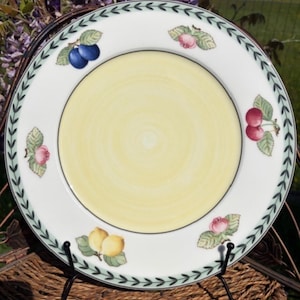 Può includere: Un piatto in ceramica bianca con un centro giallo e un bordo decorativo. Il bordo presenta un motivo a foglie verdi e illustrazioni di frutta, tra cui ciliegie, prugne e limoni. Il piatto è esposto su un supporto in metallo nero.