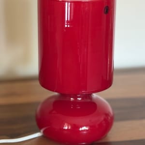 Può includere: Una lampada da tavolo in vetro rosso brillante con paralume cilindrico su una base arrotondata e bombata. L'interno dell'apertura superiore rivela un rivestimento rosa pallido e un piccolo adesivo nero circolare è visibile sul lato del paralume.