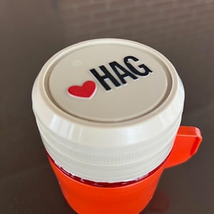 Peut inclure: Un mug orange vif avec un couvercle beige orné d'une icône de cœur rouge et du texte noir HAG imprimé sur la surface supérieure. Le mug possède une anse simple sur le côté et une texture nervurée autour du couvercle circulaire, vu du dessus.