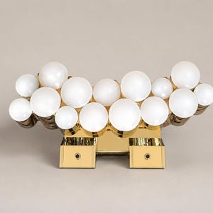 Op de afbeelding: Een wandlamp met een gepolijste goudkleurige metalen basis en een cluster van meerdere bolvormige witte glazen bollen die in een horizontale, wolkachtige formatie zijn gerangschikt. De armatuur bevat twee rechthoekige montagebeugels.