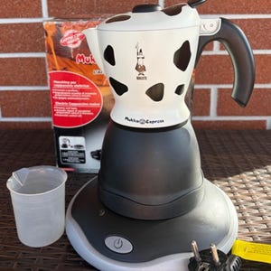 Pode incluir: Uma máquina de cappuccino elétrica branca e preta com padrão de manchas de vaca, pega e base pretas, com o logótipo Bialetti e o texto Mukka Express. O conjunto inclui um copo medidor de plástico transparente e um cabo de alimentação.