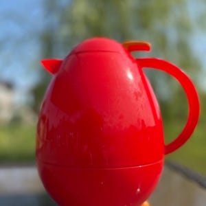 Può includere: Una caraffa termica rosso brillante a forma di uovo con una finitura liscia e lucida. Presenta un manico curvo, un piccolo beccuccio e un coperchio con un dettaglio arancione. La base è sostenuta da tre piccoli piedini rotondi di colore arancione.