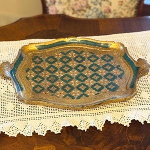 Pode incluir: Uma bandeja de servir antiga com um esquema de cores dourado e verde-azulado. A bandeja apresenta um padrão geométrico detalhado em verde-azulado e dourado, com acabamentos e alças douradas ornamentadas. Está colocada sobre uma toalha de mesa de renda branca. Dimensões: aprox. 40 cm x 30 cm.