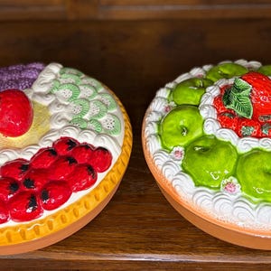 Pode incluir: Dois pratos de cerâmica em forma de torta com desenhos de frutas. Um apresenta uma variedade de frutas, incluindo bagas vermelhas, fatias de limão e um morango. O outro tem maçãs verdes e um morango. Ambos têm cobertura branca e uma base marrom.