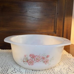 May include: White, round serving bowl with two handles. The bowl features a floral design with pink blossoms and green leaves. The bowl is sitting on a white lace tablecloth. The background is a dark wooden cabinet.