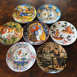 Może przedstawiać: Kolekcja siedmiu okrągłych talerzy ceramicznych z różnorodnymi ilustracjami dekoracyjnymi, w tym portretem postaci w hełmie, sceną kanału z gondolą, motywami kwiatowymi oraz scenami krajobrazowymi z postaciami w żywych kolorach.