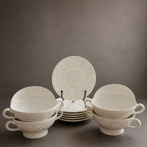 Può includere: Un set di ciotole da zuppa e piattini abbinati in ceramica color crema con un motivo floreale in rilievo. Le ciotole sono dotate di due manici laterali e una base a piedistallo, mentre i piattini presentano un bordo decorativo.