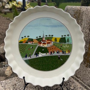 Może przedstawiać: Okrągła, biała ceramiczna forma do tarty z falistym brzegiem, ozdobiona centralną ilustracją wiejskiej scenerii. Obraz przedstawia orszak ślubny idący ścieżką przez zielone pola, sady i domy pod jasnoniebieskim niebem.