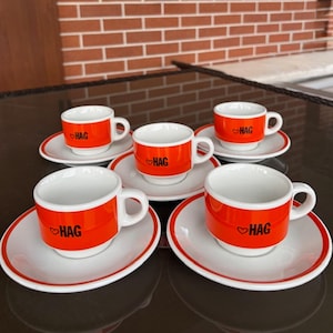 May include: A set of four white ceramic espresso cups and matching saucers, each featuring a vibrant orange band around the center. The cups are printed with the black text HAG accompanied by a small heart symbol on the orange section.