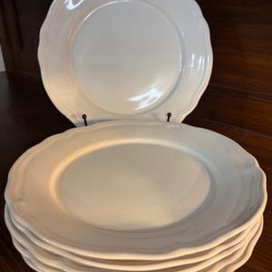 Può includere: Una pila di cinque piatti piani in ceramica bianca con una finitura liscia e lucida e un bordo smerlato ondulato. Un piatto è posizionato verticalmente su un piccolo supporto in metallo nero, mentre gli altri quattro sono impilati sotto.