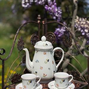 Puede incluir: Una tetera de porcelana blanca y dos tazas de té con diseños florales, colocadas sobre una superficie de mimbre marrón con un soporte de metal.