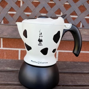 Può includere: Una caffettiera Mukka Express bianca e nera con un motivo a mucca. La caffettiera ha una base, un manico e un coperchio neri e presenta il logo Bialetti e la scritta "Mukka Express".