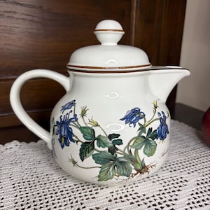 Può includere: Teiera in ceramica bianca con coperchio e manico. La teiera è decorata con un motivo floreale con fiori blu e foglie verdi. Il bordo e il coperchio sono rifiniti in marrone. La teiera è appoggiata su una tovaglia di pizzo bianco.
