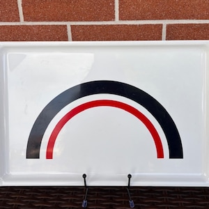 Può includere: Un vassoio da portata rettangolare bianco con un grafico centrale a forma di arcobaleno. Il design è composto da una spessa banda esterna nera, una banda centrale rossa più sottile e una spessa banda interna nera su fondo bianco.
