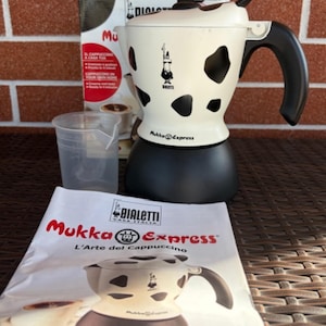 Pode incluir: Uma cafeteira de cappuccino Bialetti Mukka Express com padrão de manchas de vaca em preto e branco, base preta e alça preta. O conjunto inclui um copo medidor de plástico transparente, a caixa original e um manual de instruções.