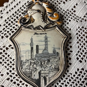 May include: A ceramic wall plaque featuring a shield shape with a depiction of Siena, Italy. The plaque has a decorative border and a helmeted figure at the top. The word "Siena" is written across the top of the shield.