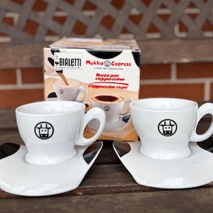 Op de afbeelding: Twee witte keramische cappuccinokopjes met bijpassende schoteltjes voorzien van een zwart koeienkop-logo. De set staat voor een Bialetti Mukka Express-doos met de tekst L'Arte del Cappuccino, Tazze per cappuccino en Cappuccino cups.