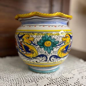 May include: A ceramic vase with a ruffled yellow rim, blue and yellow accents, and a turquoise base. The vase features a floral design with two yellow dragon-like creatures and a central teal flower.