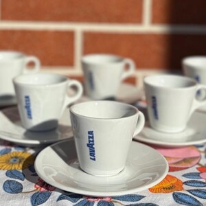 Könnte beinhalten: Ein Set aus fünf weißen Espresso-Tassen aus Keramik mit passenden Untertassen, die jeweils das blaue Lavazza-Logo auf der Vorderseite tragen. Die Tassen haben eine konische Form mit einem kleinen Henkel und die Untertassen sind rund.