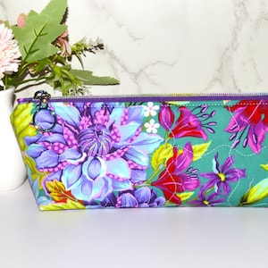 May include: A rectangular fabric zipper pouch featuring a vibrant floral pattern with large purple and blue dahlia-like flowers, smaller pink blossoms, and green leaves on a teal background. The pouch has a purple zipper and a silver metal ring pull.