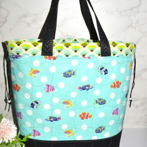 May include: A quilted tote bag featuring a light blue background with white polka dots and various colorful tropical fish. The bag has a black base, black fabric handles, and a decorative green and blue scalloped trim along the top edge.