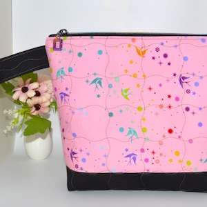 May include: Pink fabric pouch with a black base and handle. The pouch features a pattern of colorful birds, stars, and dots. A black zipper and a small purple zipper pull complete the design. The pouch is approximately 10 inches wide.