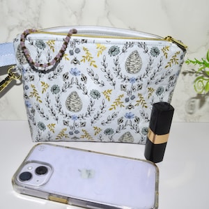 May include: A white cosmetic bag with a floral and bee pattern, a blue strap, and a gold zipper. A phone, lipstick, and a beaded bracelet are next to the bag. The bag is approximately 8 inches wide.