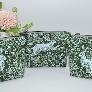May include: Three green zippered pouches with a white rabbit and floral pattern. The pouches have a silver zipper and are arranged in a row. A vase of pink flowers is in the background.