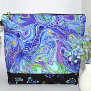 May include: A quilted fabric pouch with a swirling blue, purple, and green pattern. The pouch has a black base with butterfly accents and a gold zipper. A matching wrist strap and a small star charm are attached.