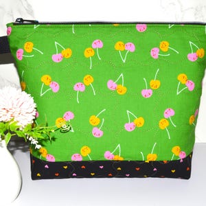 May include: A green quilted fabric bag with a black zipper and strap. The bag features a pattern of pink and yellow cherries on a green background. The bottom of the bag is black with a pattern of small hearts.