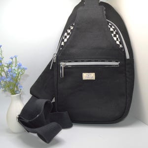 May include: Black sling bag with a checkered pattern on the side and a silver zipper. The bag has a front pocket and a black strap. A small white vase with blue flowers is to the left of the bag.