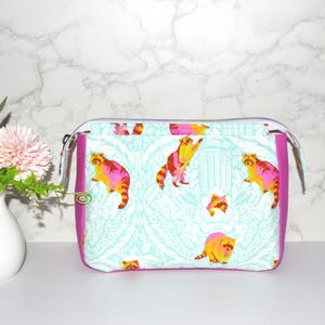 May include: A rectangular zippered pouch featuring a light blue and white damask-style background pattern with repeating illustrations of raccoons in shades of pink, orange, and yellow. The pouch has solid magenta side panels and a silver zipper.