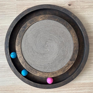 Op de afbeelding: Een ronde, donkerbruine kattenspeelgoed met een centrale kartonnen krabpaal en drie gekleurde ballen. Het speelgoed heeft een cirkelvormig ontwerp met concentrische ringen, en de ballen zijn blauw en roze.
