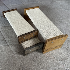 Op de afbeelding: Twee houten krabpalen voor katten met een lichtgekleurd kraboppervlak van touw. De houten frames zijn lichtbruin met decoratieve geëtste patronen. De krabpalen zijn schuin, met één iets verhoogd.