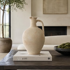 May include: A beige, textured ceramic pitcher with a rounded body, a narrow neck, and a single handle, resting on a light-colored book titled AD at 100. The pitcher has a matte, earthy finish and a simple, functional design with a small pouring spout.