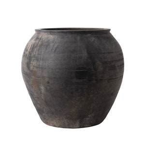 May include: A large, round, black clay pot with a wide opening. The pot has a rustic, aged appearance with visible lines and texture.