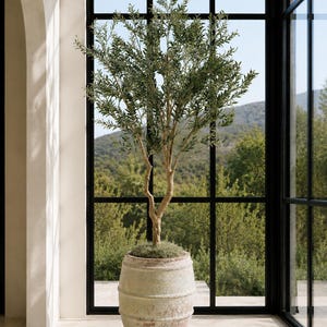 May include: A tall, slender artificial olive tree with green leaves and a textured, light-colored trunk, planted in a large, weathered, barrel-shaped ceramic planter with a distressed, earthy finish and visible horizontal ridges.